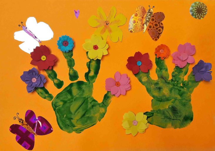 zielone dłonie namalowane farbą na pomarańczowej kartce papieru pośród kwiatów i motyli z papieru