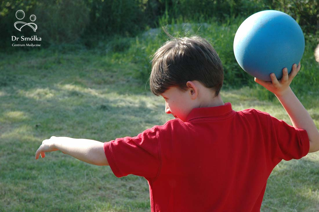 chłopiec w czerwonej koszulce rzuca niebieską piłką fizjoterapeutyczną