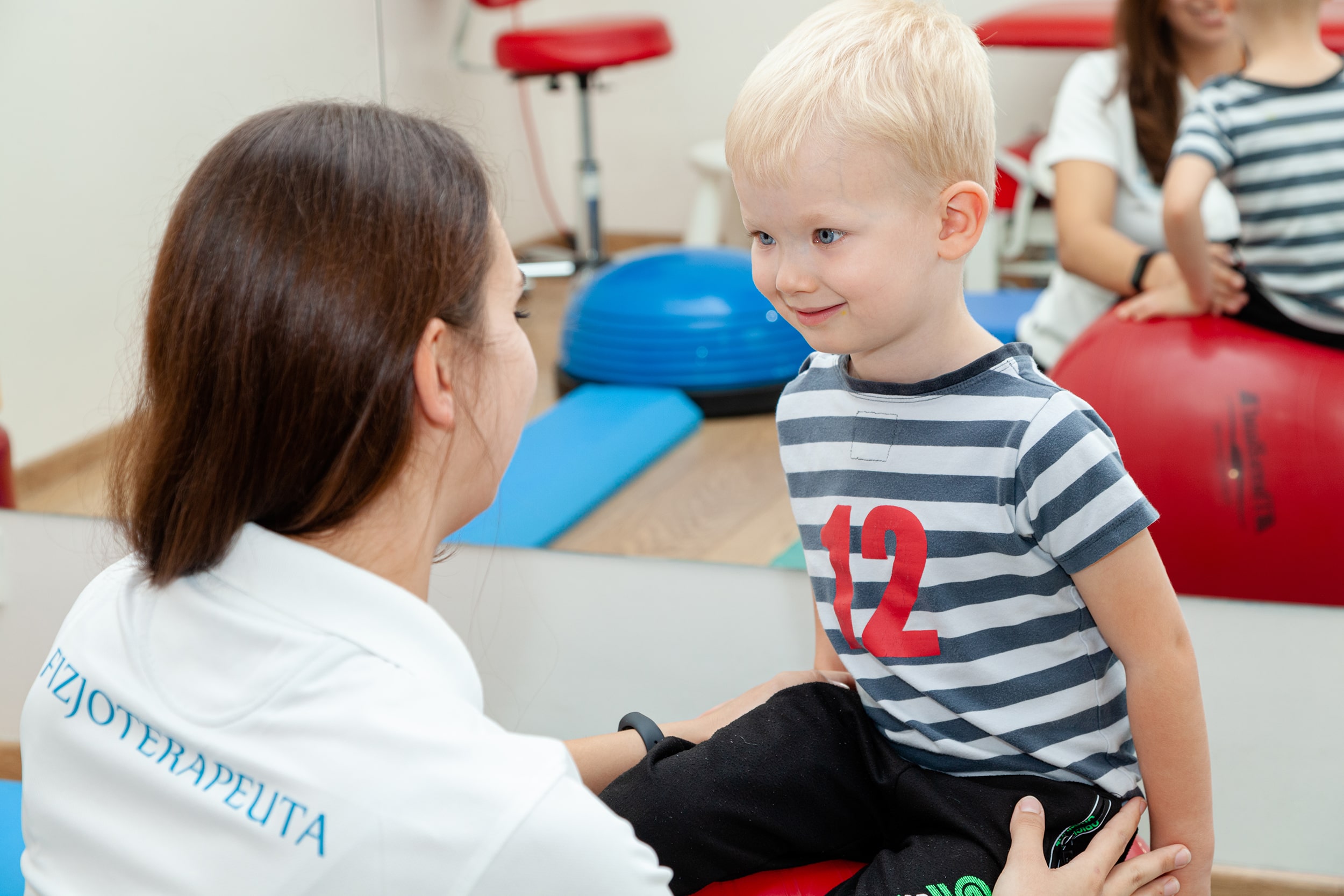 uśmiechnięte dziecko siedzi na piłce do ćwiczeń podczas rehabilitacji z fizjoterapeutą