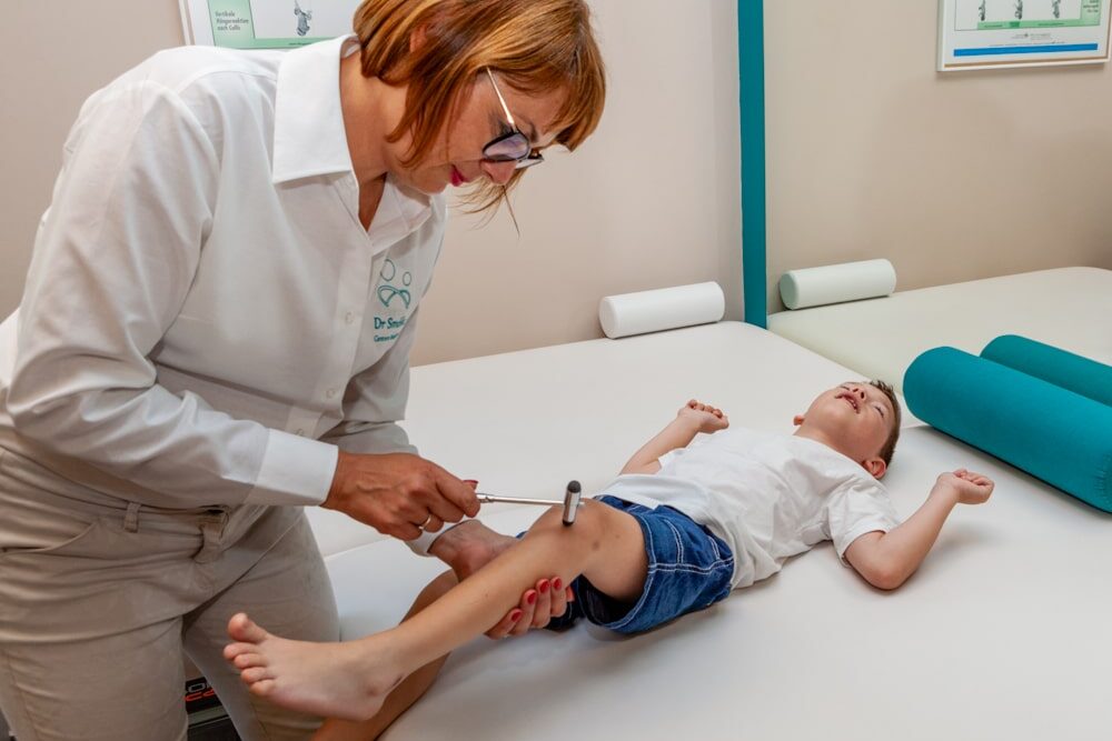 lekarz podczas badania odruchów fizjologicznych uderza młoteczkiem w kolano leżącego dziecka