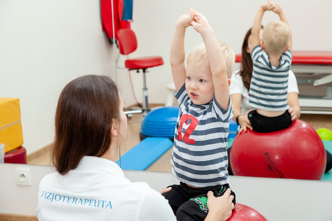dziecko wykonuje ćwiczenie na piłce gimnastycznej podczas rehabilitacji na sali z fizjoterapeutą